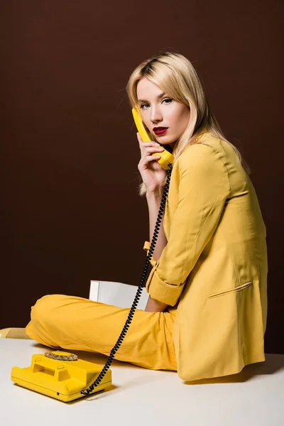Приваблива стильна блондинка в жовтому одязі розмовляє роторним телефоном і дивиться на камеру на коричневому — стокове фото