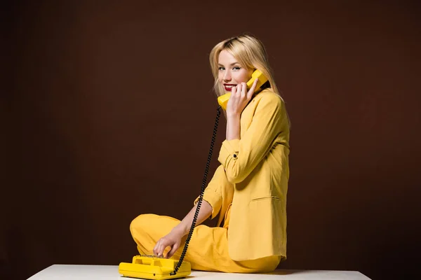 Beautiful young woman talking by yellow vintage phone and smiling at camera on brown — Stock Photo