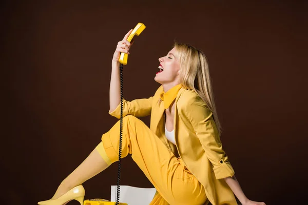 Excitada chica rubia elegante sosteniendo el auricular amarillo y sentado en marrón - foto de stock