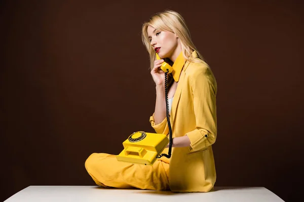 Beautiful stylish blonde girl talking by yellow vintage phone and looking away on brown — Stock Photo