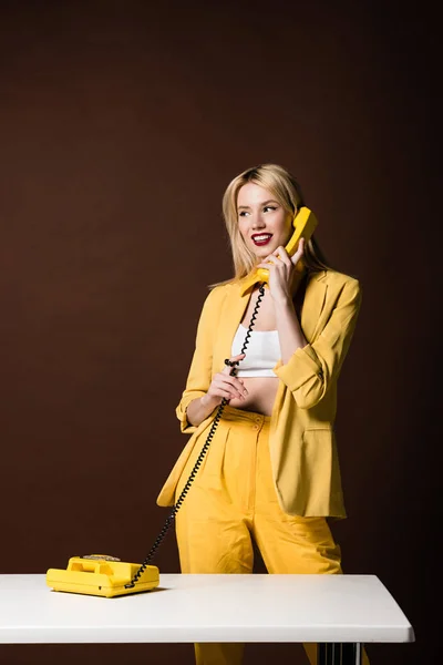 Beautiful stylish blonde woman talking by yellow vintage phone and looking away on brown — Stock Photo