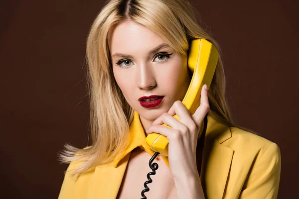 Привлекательная блондинка разговаривает по желтому винтажному телефону и смотрит на камеру, изолированную на коричневый — стоковое фото