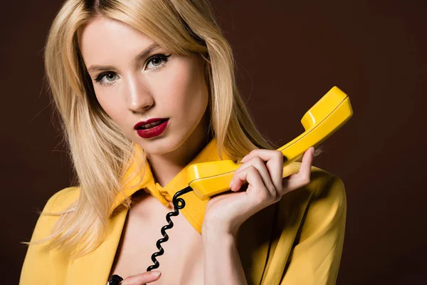 Приваблива блондинка тримає жовтий телефон і дивиться на камеру ізольовано на коричневому — стокове фото