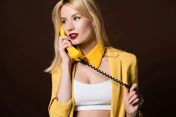 Красива стильна дівчина розмовляє жовтим старовинним телефоном і дивиться ізольовано на коричневий — стокове фото