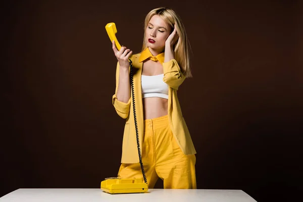 Hermosa chica rubia joven en ropa amarilla sosteniendo el teléfono en marrón - foto de stock