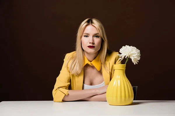 Красива стильна блондинка дивиться на камеру, сидячи за столом з білою квіткою в жовтій вазі на коричневому — стокове фото
