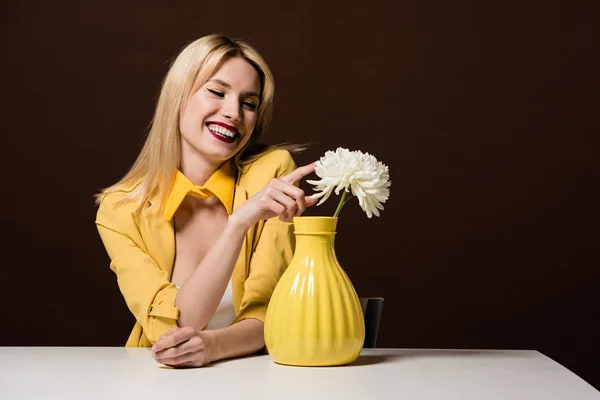 Schöne lächelnde blonde Frau in stylischem gelben Outfit, die weiße Blume in der Vase auf braun berührt — Stockfoto