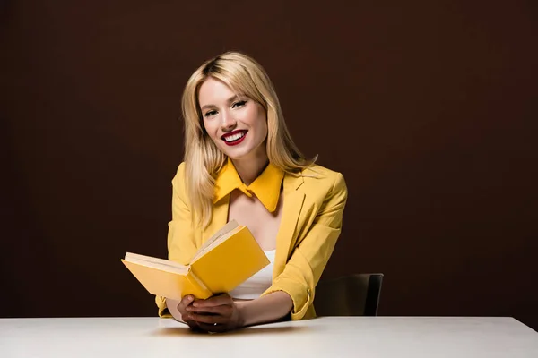 Hermosa chica rubia elegante sosteniendo libro amarillo y sonriendo a la cámara en marrón - foto de stock