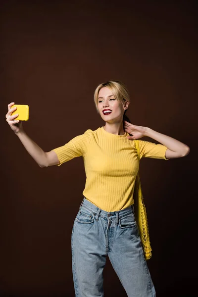 Smiling blonde woman with bananas in string bag taking selfie by yellow smartphone isolated on brown — Stock Photo