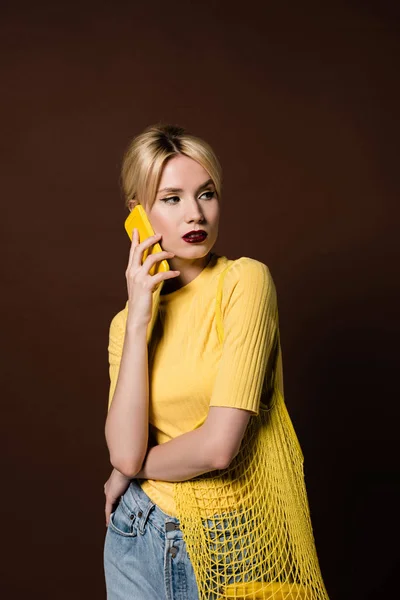 Hermosa mujer rubia con bolsa de cuerda hablando por teléfono inteligente amarillo y mirando hacia otro lado aislado en marrón - foto de stock