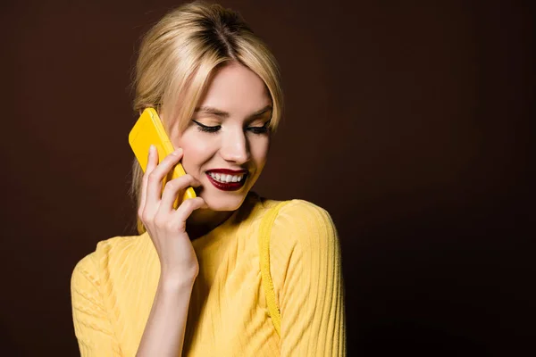 Hermosa chica rubia sonriente hablando por teléfono inteligente amarillo aislado en marrón - foto de stock