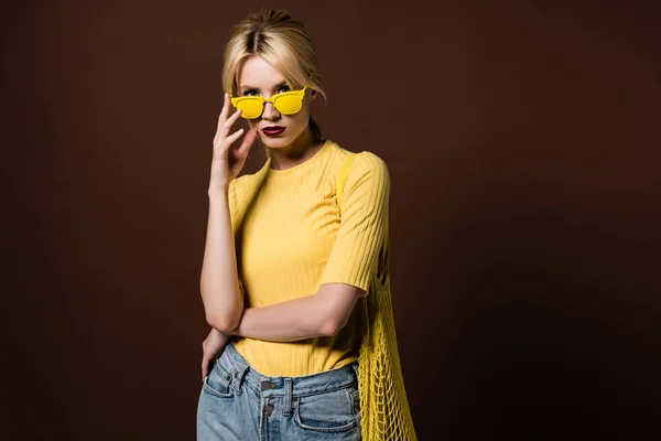 Elegante chica rubia con bolsa de hilo con gafas de sol amarillas y mirando a la cámara aislada en marrón - foto de stock