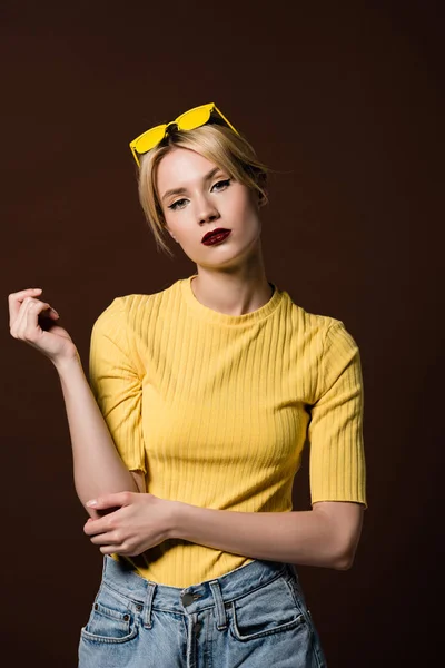 Retrato de hermosa chica rubia con estilo en gafas de sol amarillas en la cabeza mirando a la cámara aislada en marrón - foto de stock