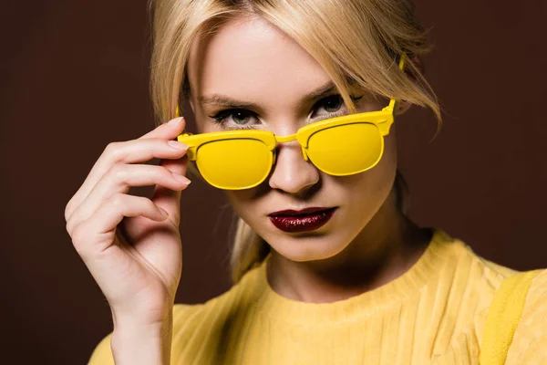 Portrait en gros plan de belle jeune femme blonde aux lunettes de soleil jaunes regardant la caméra isolée sur brun — Photo de stock