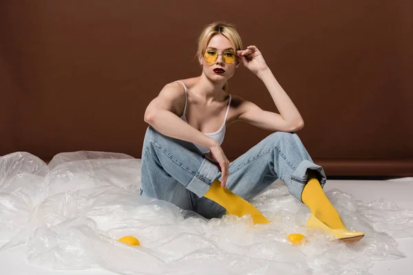 Красива стильна молода блондинка в жовтих сонцезахисних окулярах сидить і дивиться на камеру в студії — стокове фото
