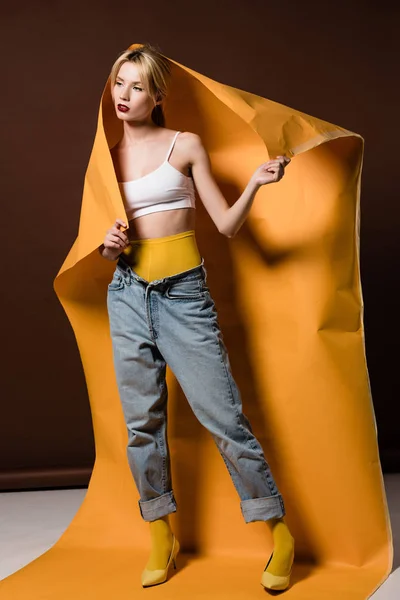 Bela menina loira elegante posando com papel laranja e olhando para longe em marrom — Fotografia de Stock