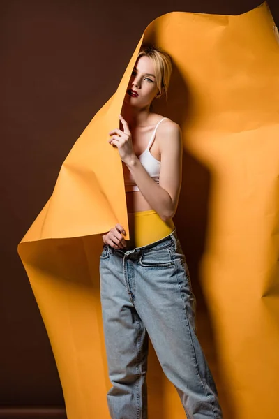 Belle fille blonde élégante posant avec du papier orange et regardant la caméra sur brun — Photo de stock