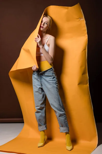 Vue pleine longueur de belle fille blonde élégante posant avec du papier orange et regardant la caméra sur brun — Photo de stock