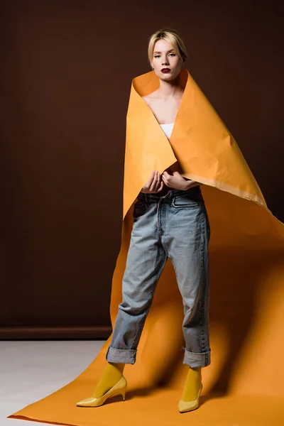 Full length view of beautiful stylish blonde girl wrapped in orange paper looking at camera on brown — Stock Photo