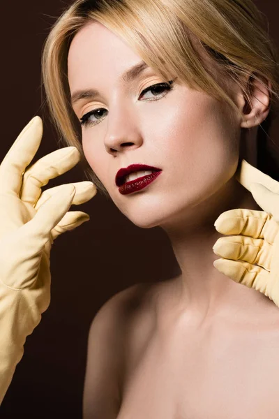 Manos humanas en guantes amarillos y hermosa chica rubia desnuda mirando a la cámara en marrón - foto de stock