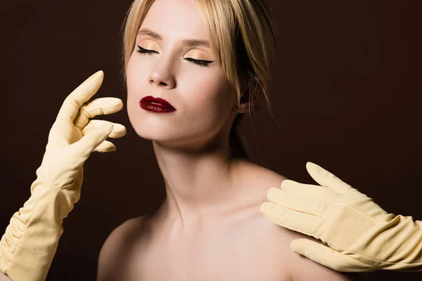 Fille blonde nue avec les yeux fermés et les mains humaines en gants jaunes sur brun — Photo de stock