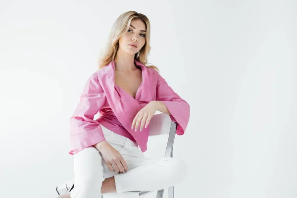 Beautiful young blond woman in stylish clothing sitting on white chair on white backdrop — Stock Photo