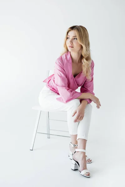 Beautiful pensive blond woman in stylish clothing sitting on white chair isolated on white — Stock Photo