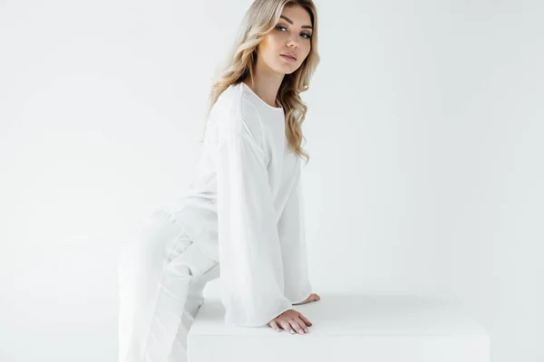 Vista lateral de la hermosa mujer rubia en ropa blanca mirando a la cámara aislada en blanco - foto de stock