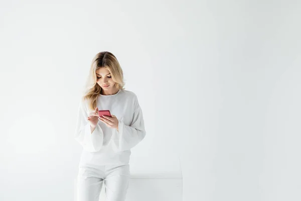 Портрет блондинки в белой одежде с помощью смартфона, изолированного на сером — стоковое фото