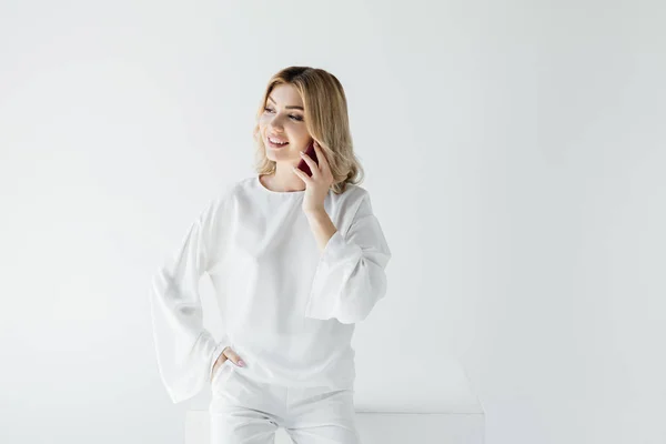 Портрет усміхненої блондинки в білому одязі, що говорить на смартфоні ізольовано на сірому — стокове фото