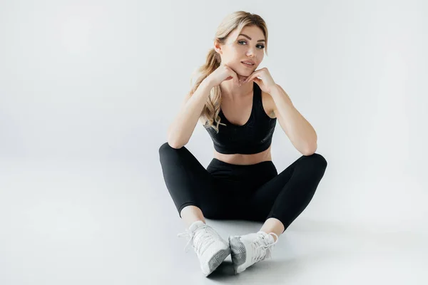 Deportista atractiva en ropa deportiva negro sentado en el suelo aislado en gris - foto de stock