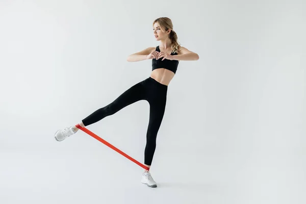 Молодая спортсменка упражняется с резиновой лентой на ногах, изолированных на сером — стоковое фото