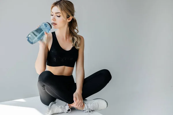 Athletische Frau in schwarzer Sportbekleidung trinkt Wasser aus Sportflasche, während sie auf einem weißen Würfel vor grauem Hintergrund sitzt — Stockfoto
