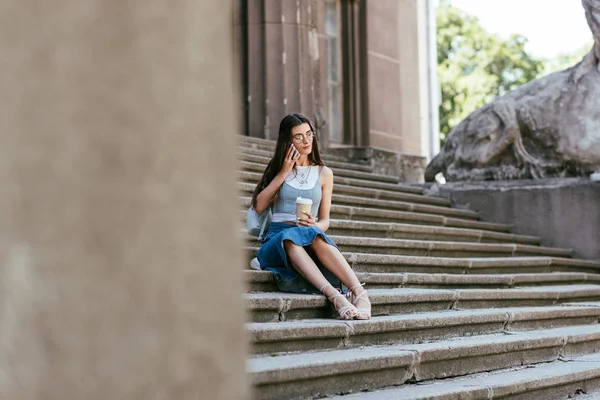 Selektiver Fokus der jungen Frau mit Kaffee, um auf Treppen zu sitzen und mit dem Smartphone zu sprechen — Stockfoto