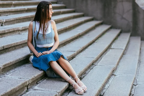 Beautiful smiling young woman in eyeglasses sitting on stairs and looking away — Stock Photo