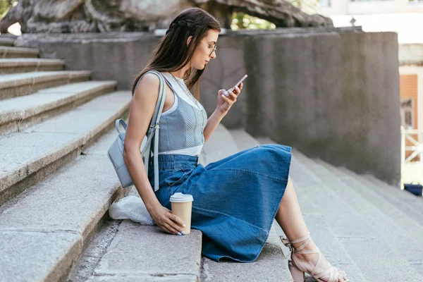 Side view of beautiful girl holding paper cup and using smartphone while sitting on stairs — Stock Photo