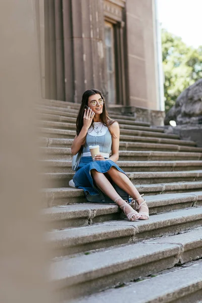 Selektiver Fokus einer jungen Frau, die Pappbecher in der Hand hält und mit dem Smartphone spricht, während sie auf der Treppe sitzt — Stockfoto