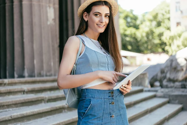 Красива молода жінка в капелюсі використовує цифровий планшет і посміхається на камеру, стоячи на сходах — стокове фото