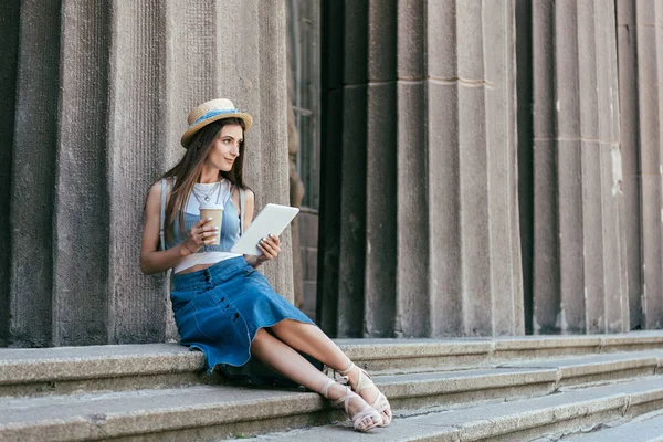 Schöne lächelnde junge Frau mit Hut, Pappbecher und digitales Tablet in der Hand, während sie auf der Treppe sitzt und wegschaut — Stockfoto