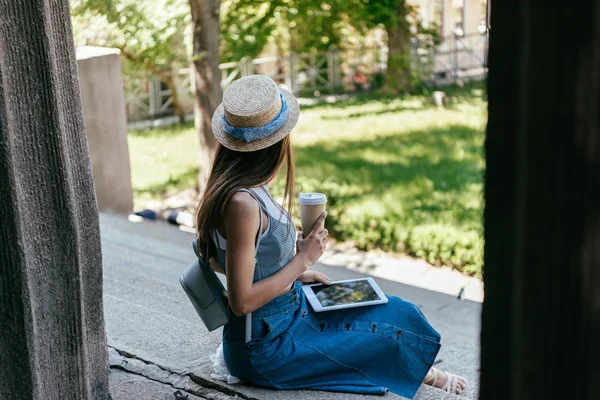 Вибірковий фокус молодої жінки, що тримає паперову чашку і використовує цифровий планшет, сидячи на сходах — стокове фото