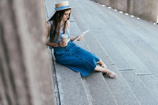 High angle view of young woman holding paper cup and using digital tablet while sitting on stairs — Stock Photo