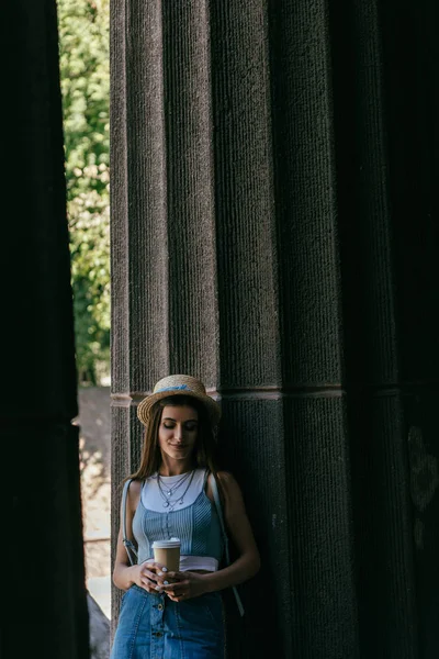 Красивая улыбающаяся молодая женщина в шляпе держит кофе, чтобы пойти и стоять между колоннами — стоковое фото