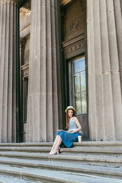 Belle fille en chapeau regardant tasse en papier tout en étant assis sur les escaliers — Photo de stock