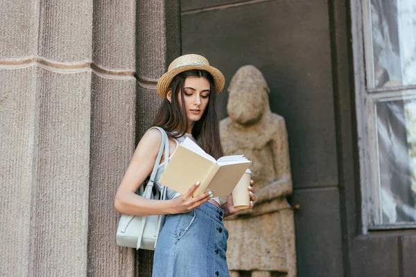 Красивая девушка в шляпе с бумажной чашкой и чтение книги возле колонны — стоковое фото