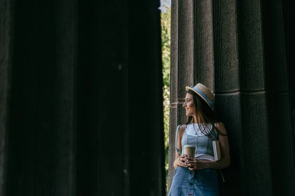 Bela jovem sorridente com livro e café para ir de pé entre colunas e olhando para longe — Fotografia de Stock