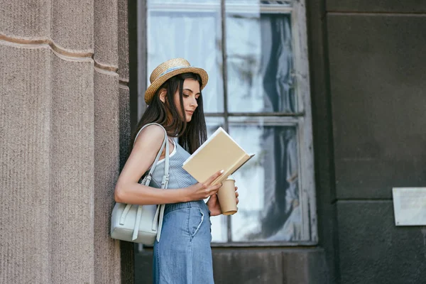 Vista lateral da menina atraente no chapéu segurando café para ir e ler livro perto da coluna — Fotografia de Stock