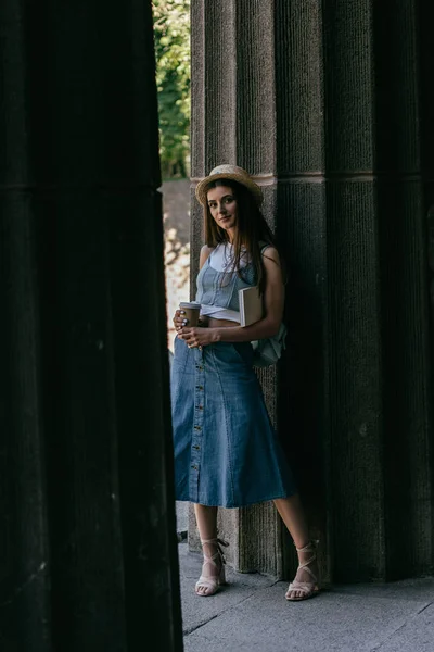Hermosa joven con libro y café para ir de pie entre columnas y mirando a la cámara - foto de stock