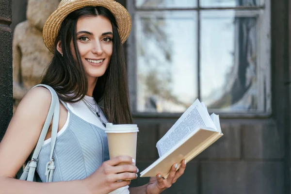 Chica atractiva con libro de lectura taza de papel y sonriendo a la cámara - foto de stock