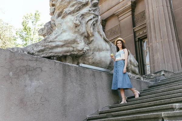 Vista ad angolo basso di giovane donna con libro e mela che cammina sulle scale e sorride alla macchina fotografica — Foto stock