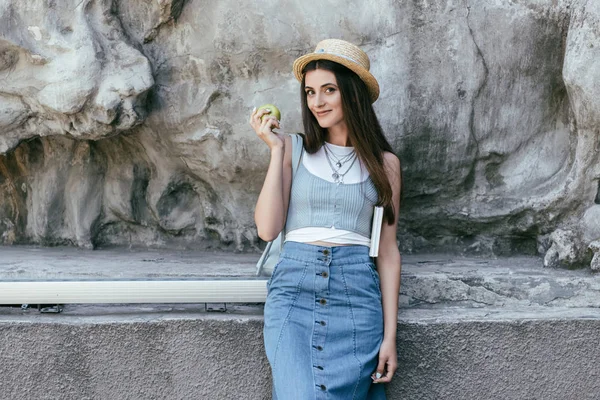 Hermosa sonriente joven en sombrero sosteniendo manzana y libro - foto de stock